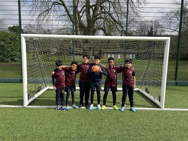 Six Long Close School pupils in maroon sports shirts stand in front of a football goal on an artificial pitch, one holding a ball.